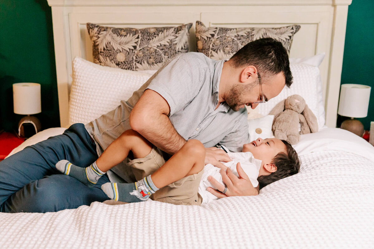 Young dad tickles his 5 year old son on the parent's bed, surrounded by white blankets. They're both laughing.