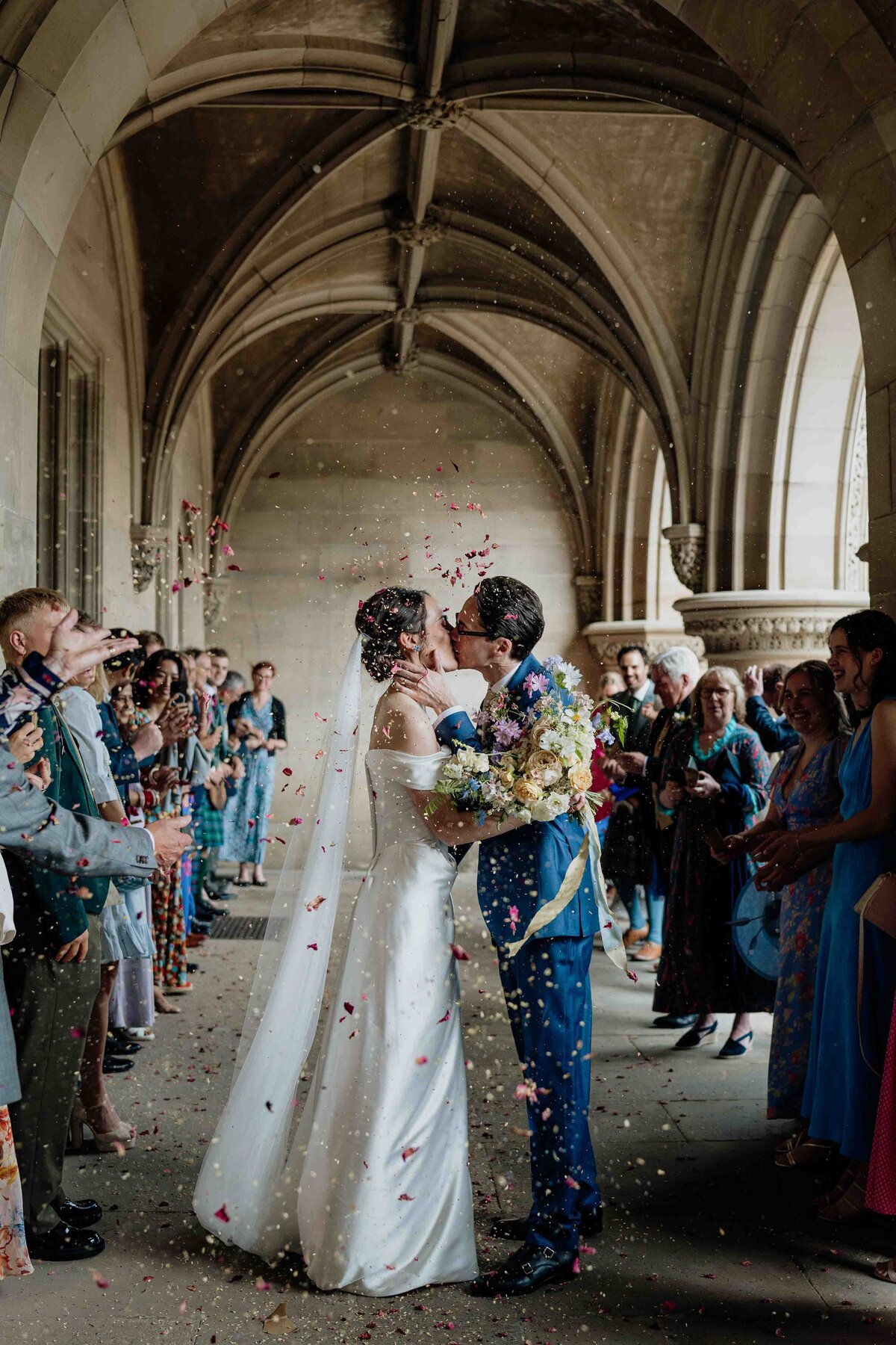 A newlywed couple are photographed sharing a kiss under an arched walkway at Fettes College in Edinburgh, surrounded by wedding guests tossing flower petals. The bride dazzles in a white dress and veil with a bouquet, while the groom looks dapper in his dark suit. The setting is elegant and celebratory.