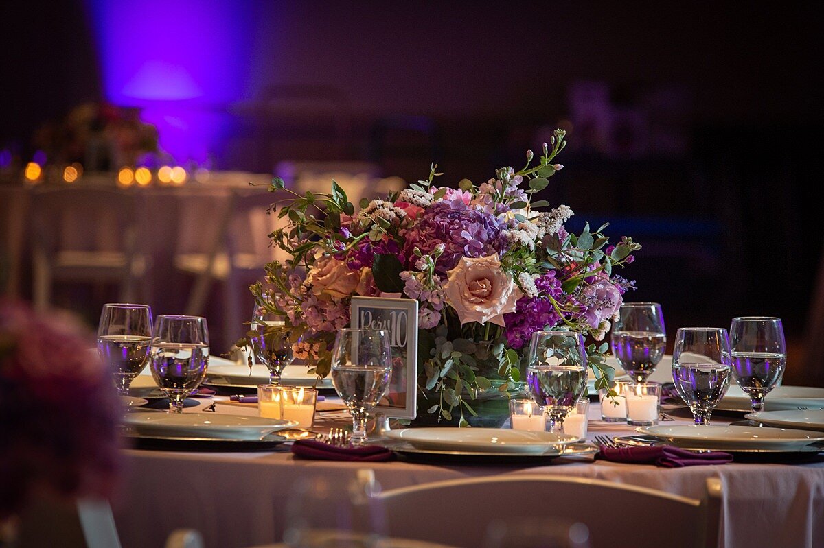 Reception tables with purple linens. The tables are set with water glasses, silver chargers with white plates and decorated with votive candles and a low centerpiece of lilac roses, purple hydrangea, and other pink and purple flowers at The Liff Center.