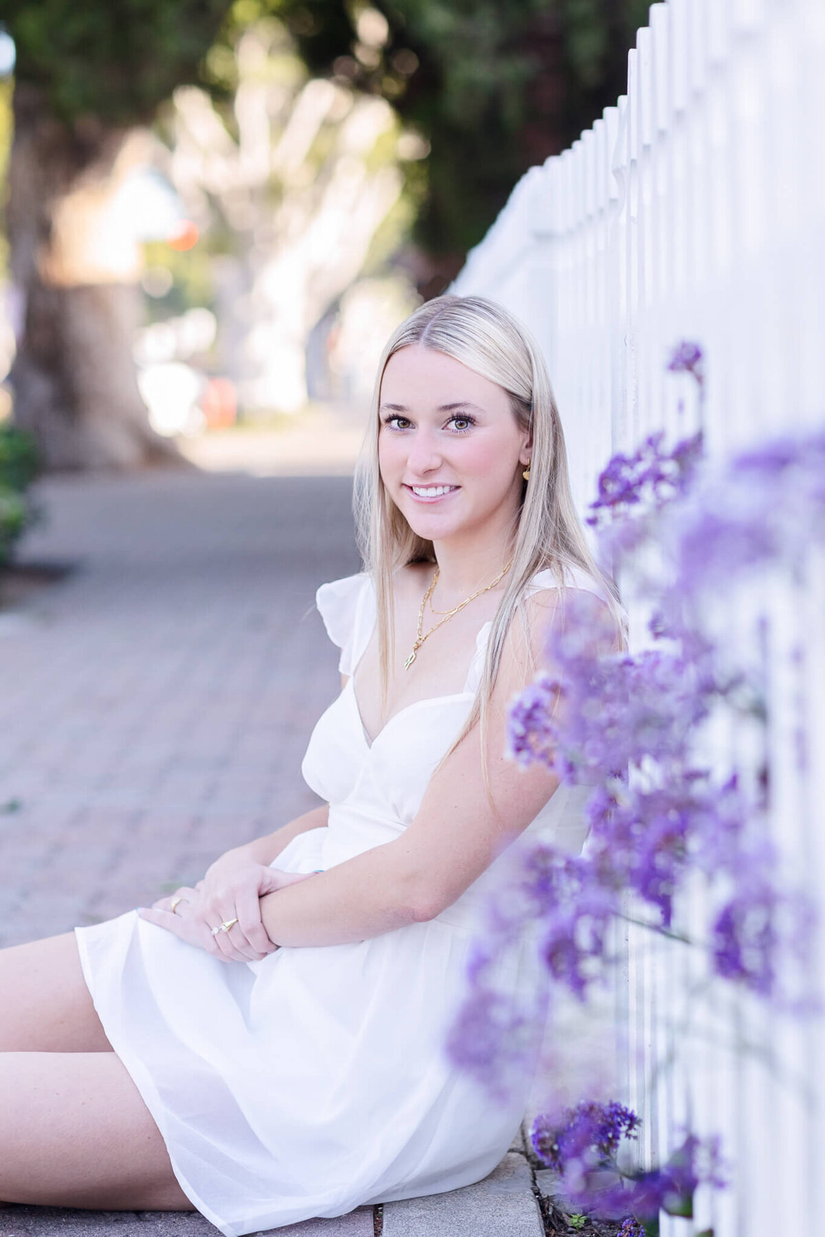 A young woman with long blonde hair sits on the ground next to a white picket fence. She is wearing a white dress and smiling slightly. Purple flowers grow along the fence, and the background is a softly blurred pathway with trees.