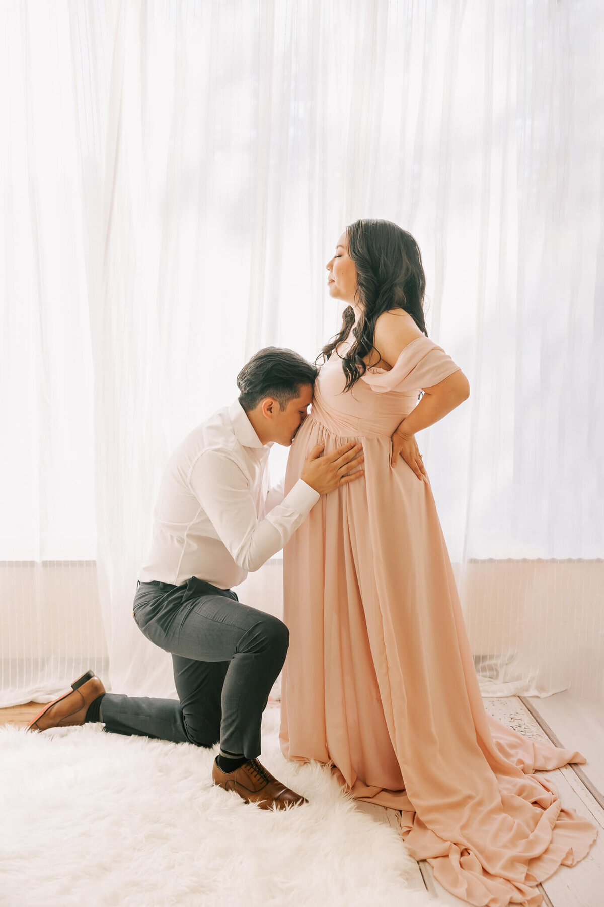 portrait of a man kissing his pregnant wife's belly in portland oregon studio maternity session. She is wearing a pink dress and he is wearing white shirt and slacks.
