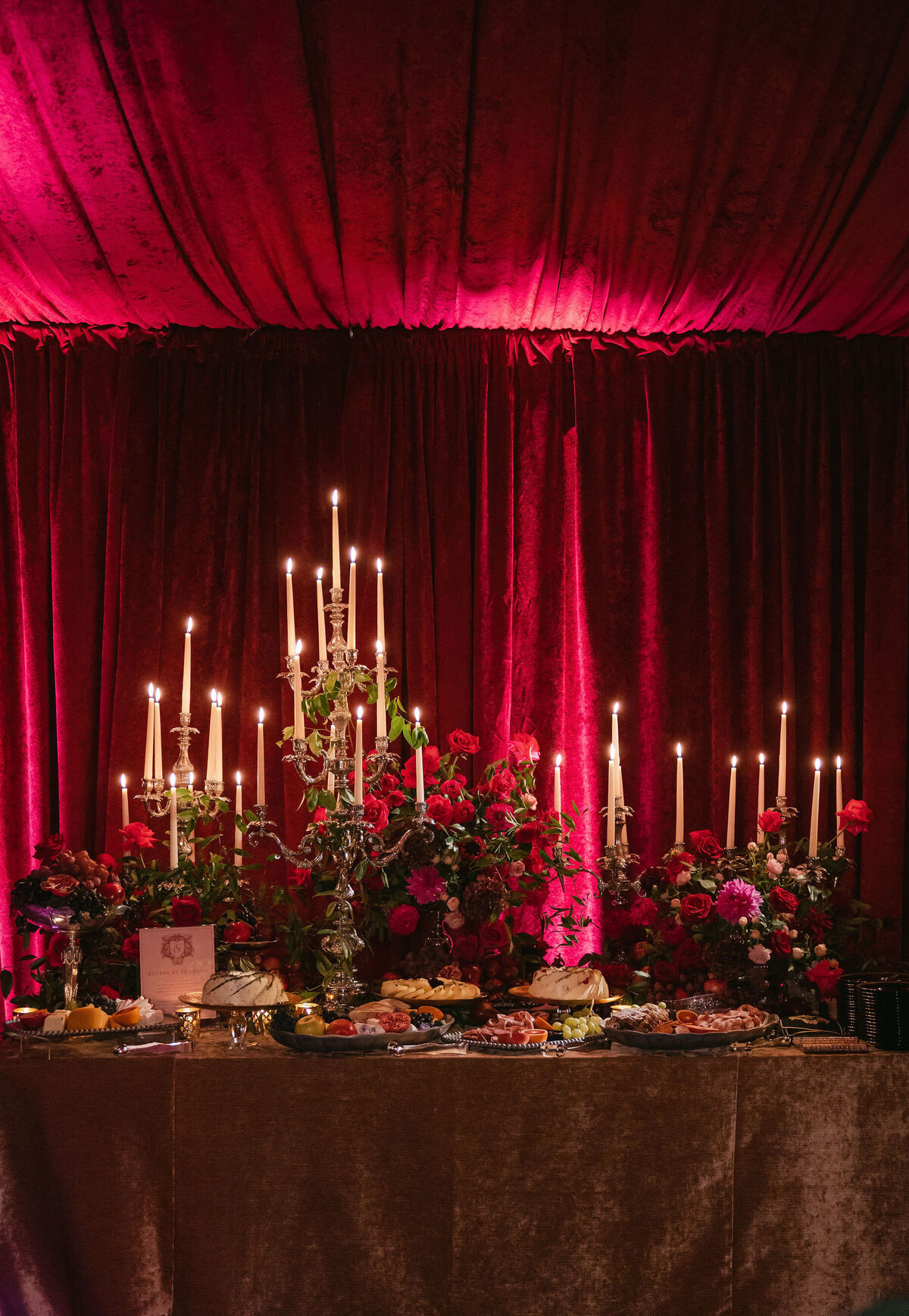 Captured by photographer Elisabetta Marzetti and videographers Clayton Films, this stunning photo was taken at a tented wedding in Virginia. A luxurious table setting steals the spotlight, showcasing an array of delectable food amidst tall lit candles and lavish floral arrangements, prominently featuring vibrant red roses. The scene is further enhanced by the presence of rich burgundy drapes in the backdrop, softly illuminated by warm, ambient lighting, exuding an opulent and romantic atmosphere.