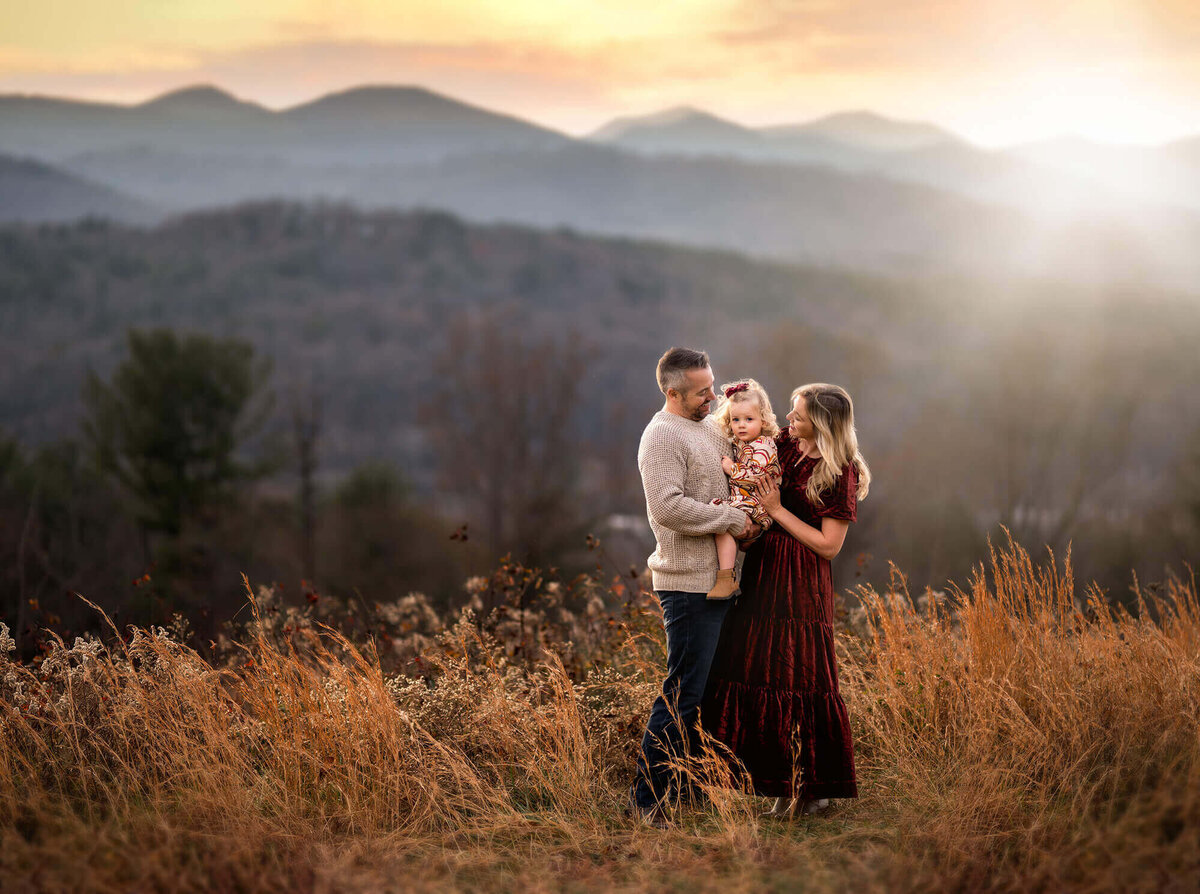 A mom, dad and daughter cuddle on a mountain top during their portrait session with an Asheville Family Photographer