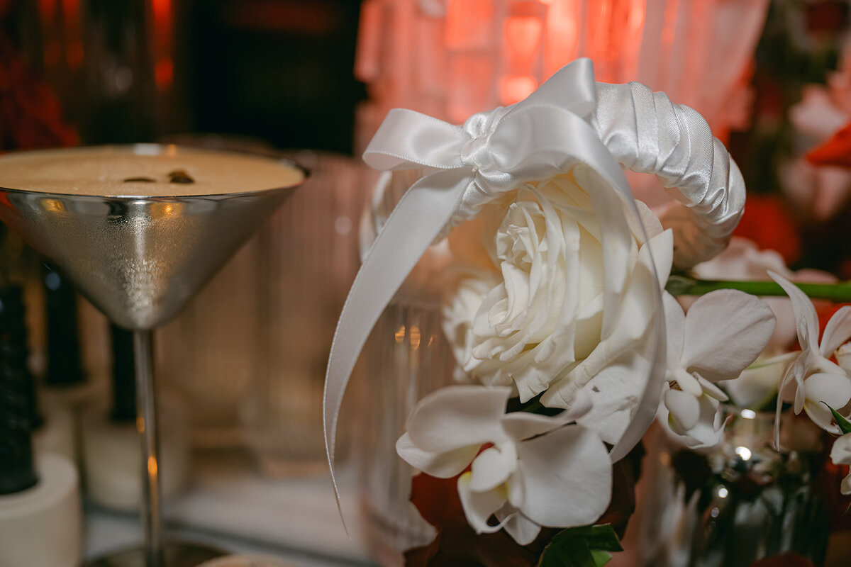 A close-up of a white rose  with silk ribbon next to a cocktail glass, styled for a luxury wedding at The Battalion in San Antonio.