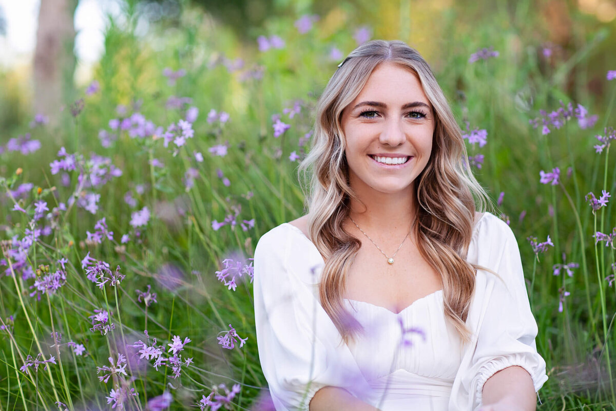 A young woman with long, wavy hair is sitting in a field of purple flowers, smiling at the camera. She is wearing a white blouse and has natural, outdoor lighting highlighting her. The background is lush and green.