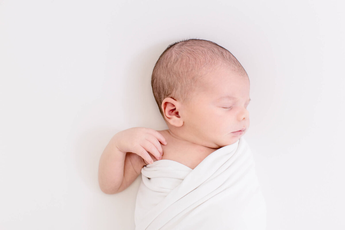 A newborn baby wrapped in a soft white blanket sleeps peacefully on a white background. The baby has closed eyes and a serene expression, lying slightly on one side.