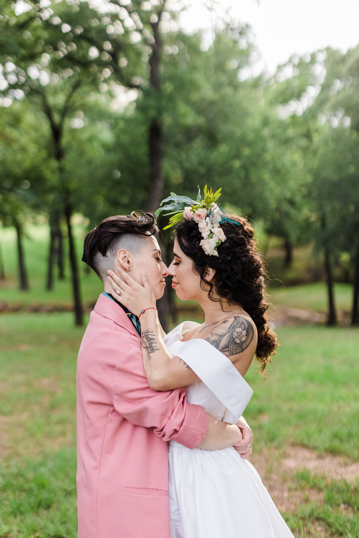 An outdoor photo of two brides holding each other close and bringing each other in for a kiss. The bride on the left is wearing a salmon suit with a colorful tie while the bride on the right is wearing an elegant white dress with a flower crown.