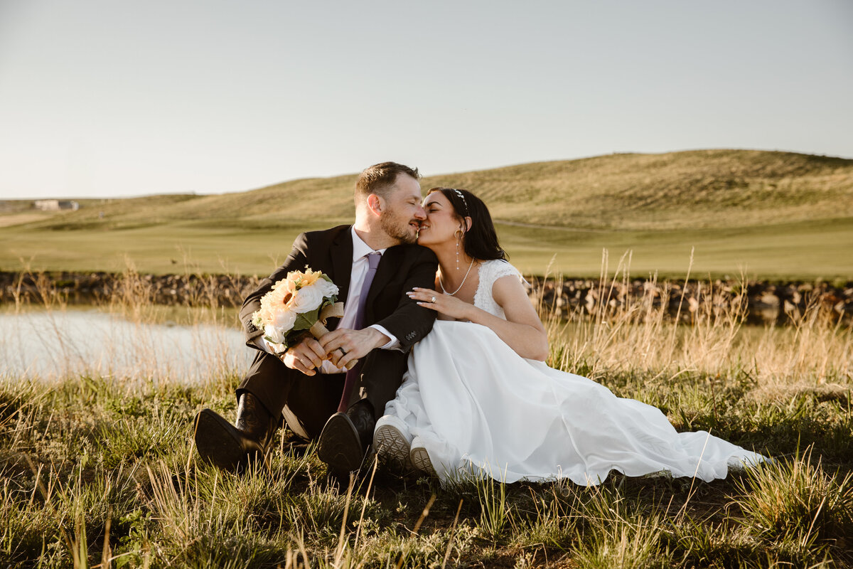 Bride and groom sitting in tall grass beside a pond at sunset, sharing a gentle kiss with rolling green hills behind them. The bride holds a bouquet and wears sneakers with her dress.