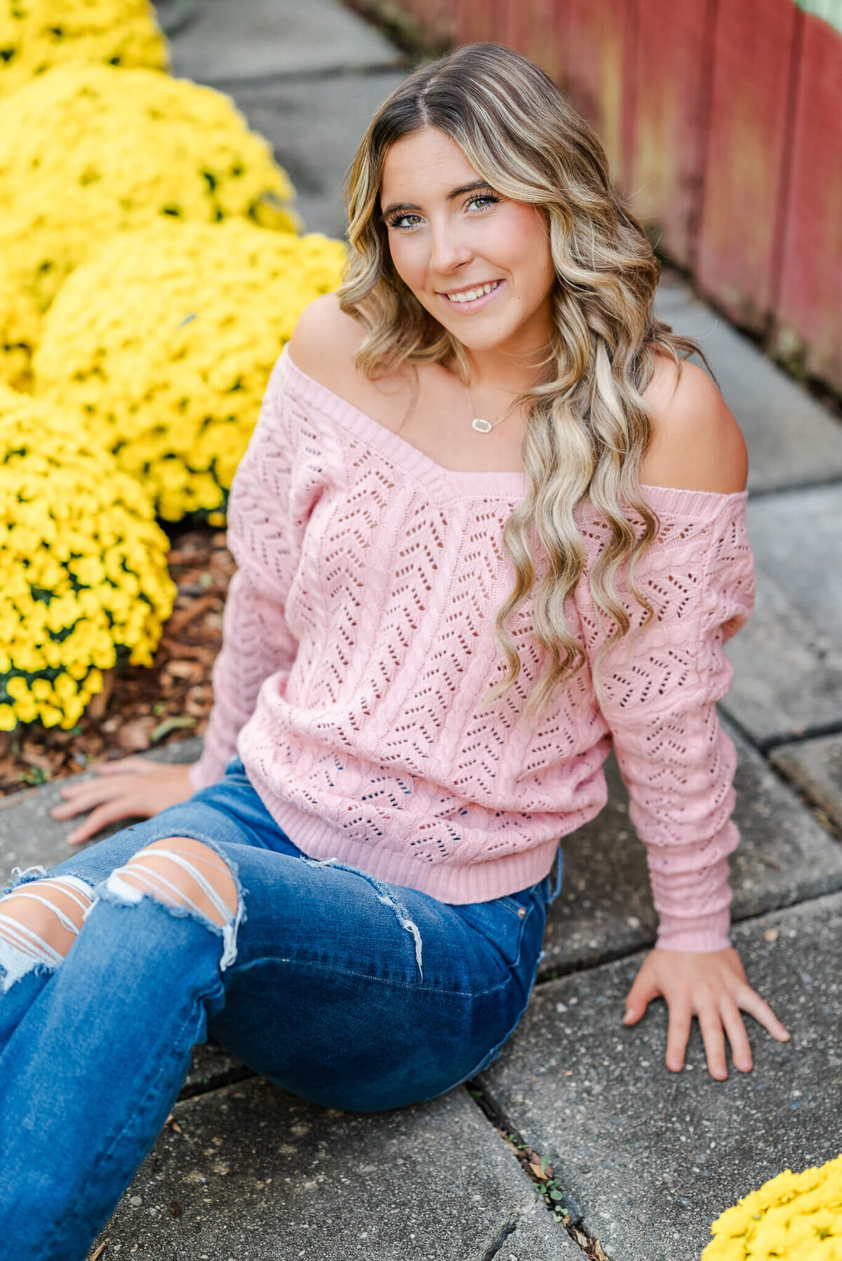 A high school senior, wearing jeans and a pink sweater, sits on some paving stones near some yellow mum bushes.