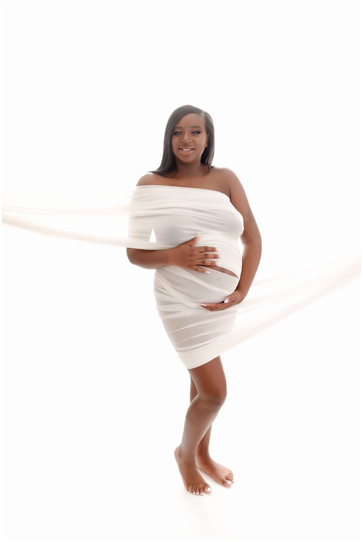 Pregnant woman wrapped in white fabric, standing on white background. She cradles her baby bump with a serene smile on her face, radiating joy and anticipation.