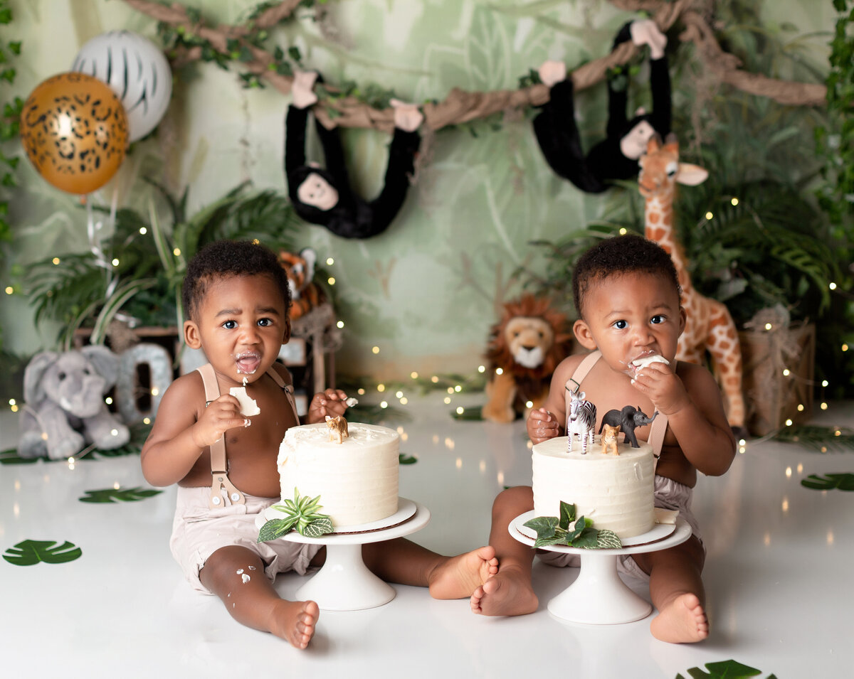 Safari jungle twin boy cake smash at West Palm Beach with top cake smash photographer. Boys have white cakes topped with safari jungle animales and greenery. Both are taking a bite of cake and looking at the camera. In the background, there is greenery, balloons, and jungle animals.