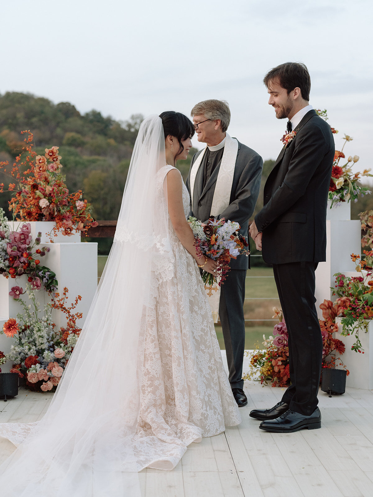 Sculptural columns wedding ceremony backdrop. Stair step white ceremony columns adorned with sculptural floral meadows in fall hues. Clear top tent build out for fall wedding reception in Tennessee hills. Wire threaded orchids, bittersweet berries, butterfly ranunculus, and tulips are highlighted in this wedding ceremony backdrop. Artistic fall color palette in copper, gold, ruby, lavender, and rust. Design by Rosemary & Finch Floral Design in Nashville, TN.