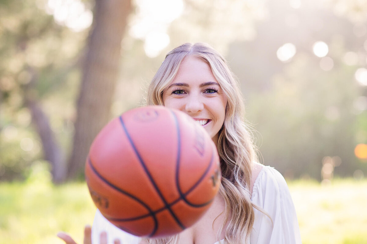 A young woman with long blonde hair smiles while holding a basketball in the foreground. The background is a sunlit park with trees and greenery, creating a cheerful and playful atmosphere.