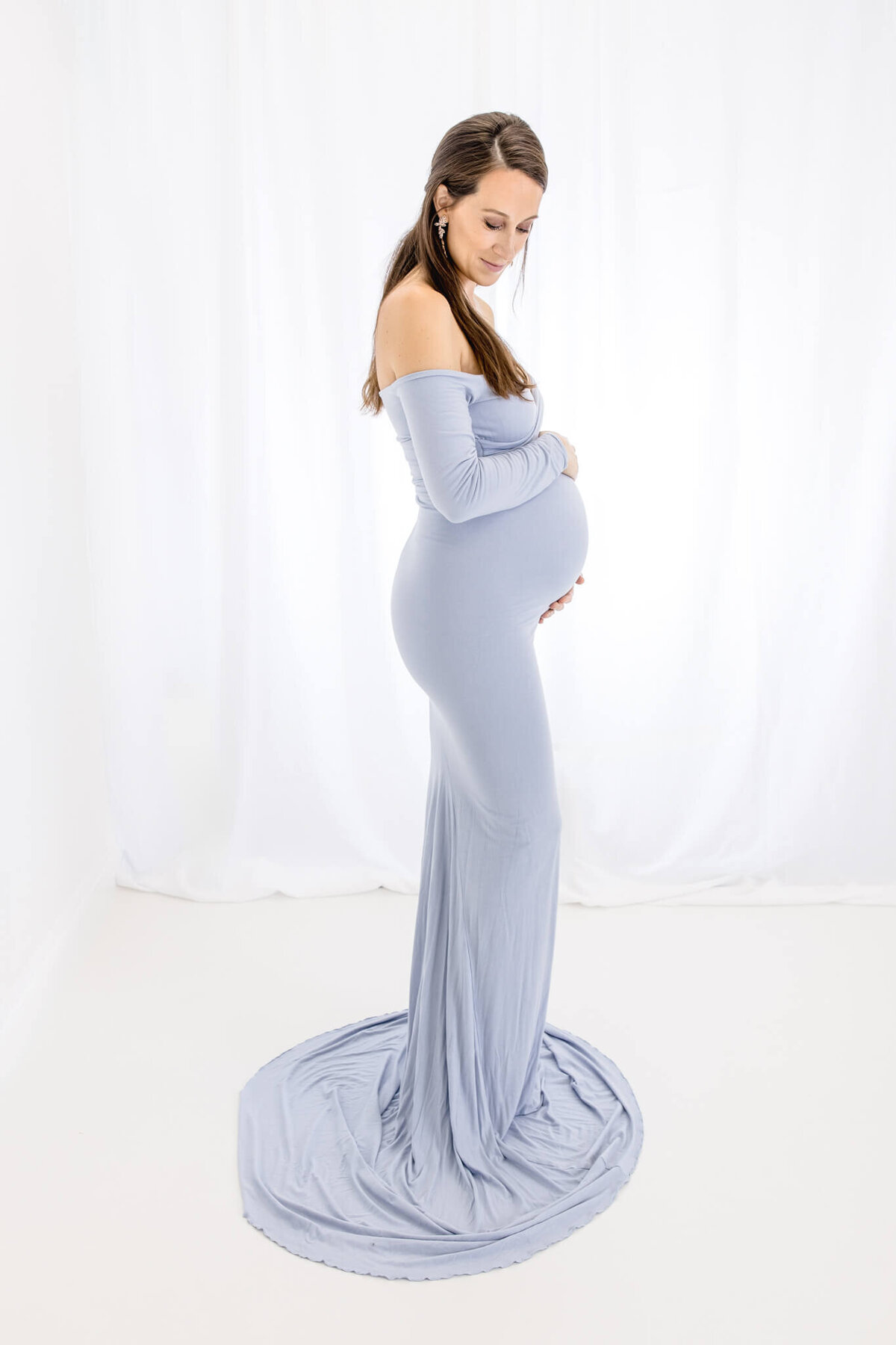 A pregnant woman in a flowing, off-shoulder light blue gown stands against a white background, gently cradling her belly and looking downward with a tranquil expression.