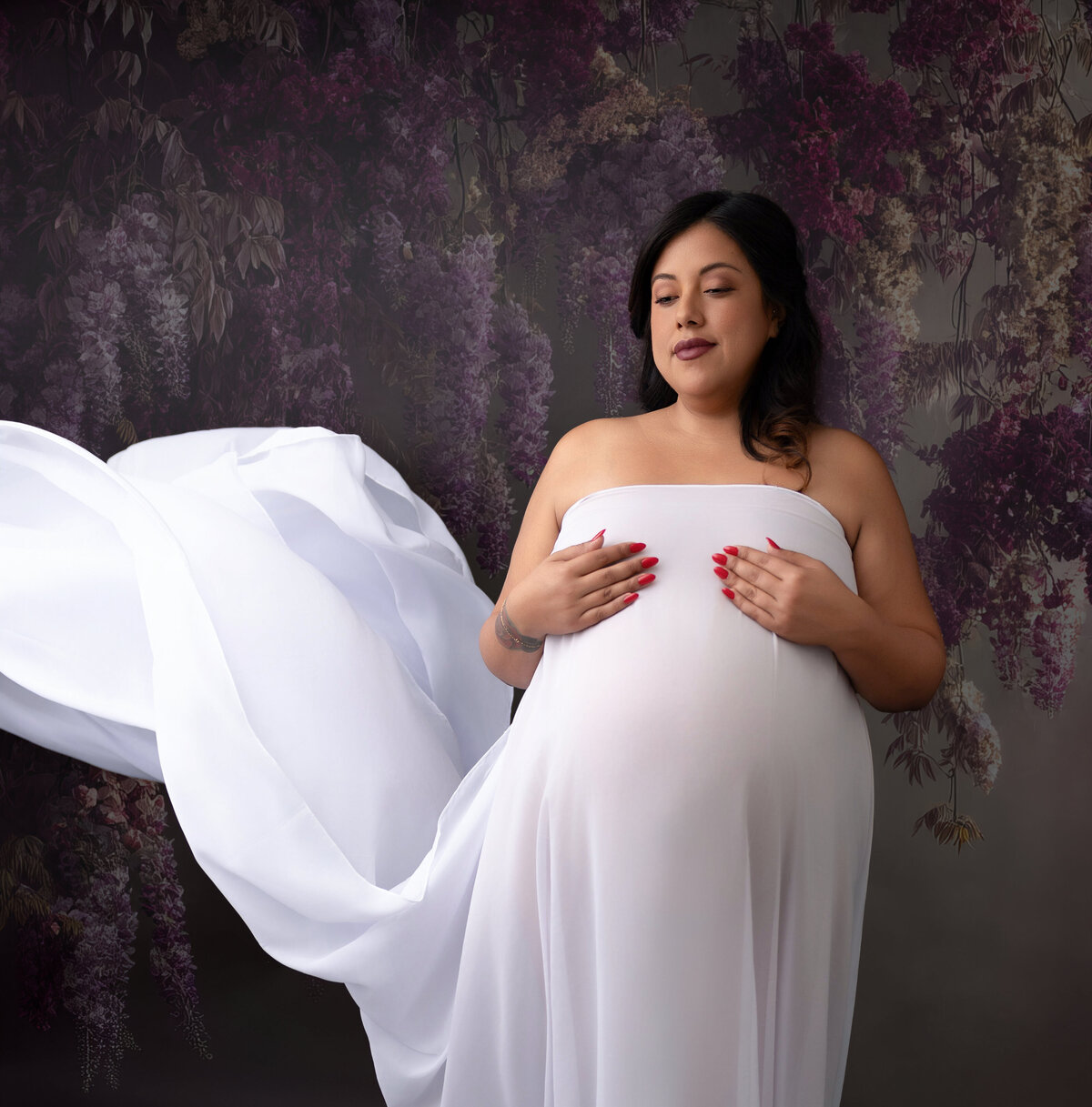 An expecting mother wearing a white silk fabric and posed against a purple color backdrop.