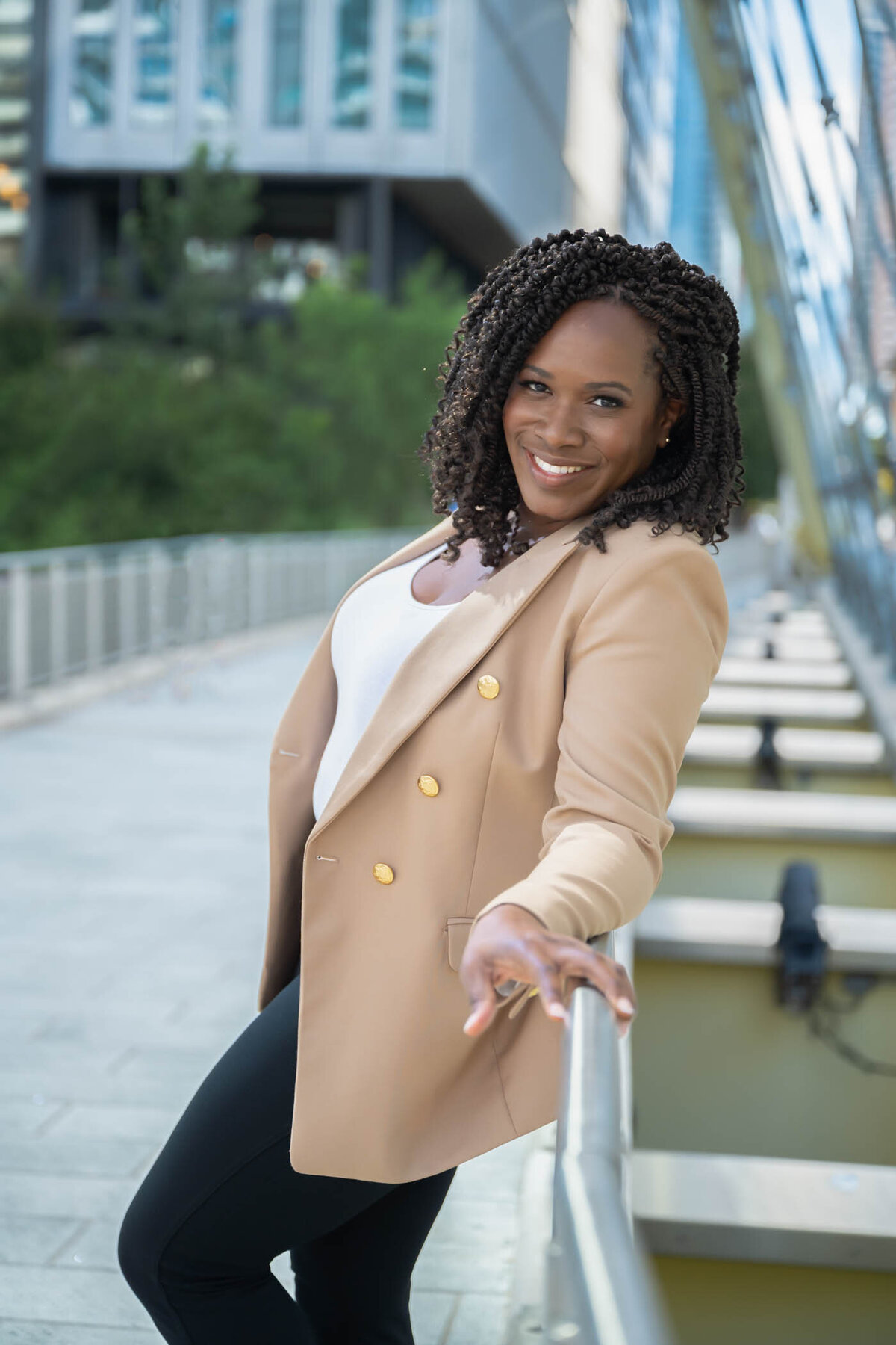 A portrait of a woman on the side of a bridge wearing a beige jacket and black pants smiling
