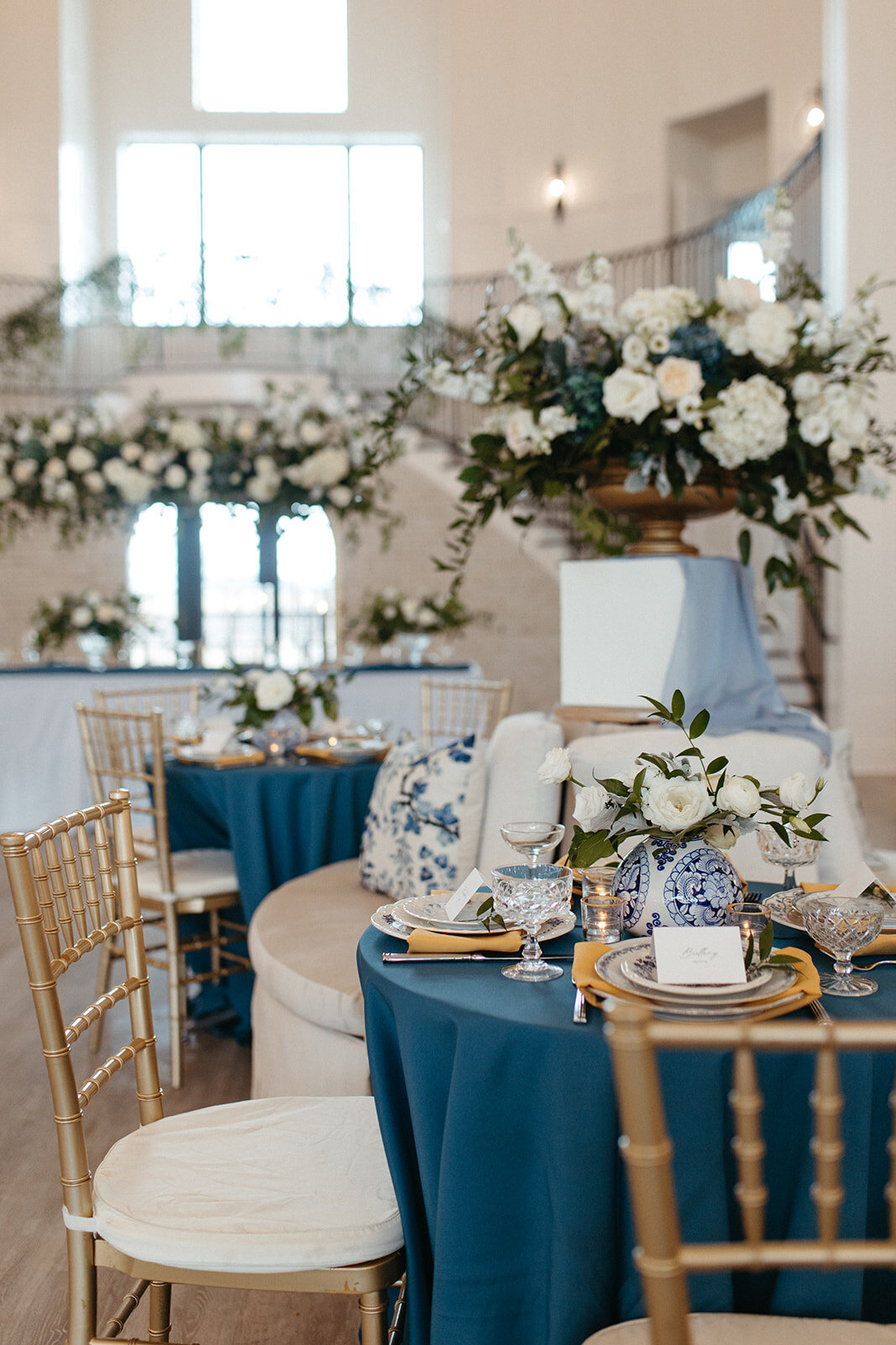 A bright and airy banquet room  with rounded tables in blue linen and gold chairs with white floral arrangements