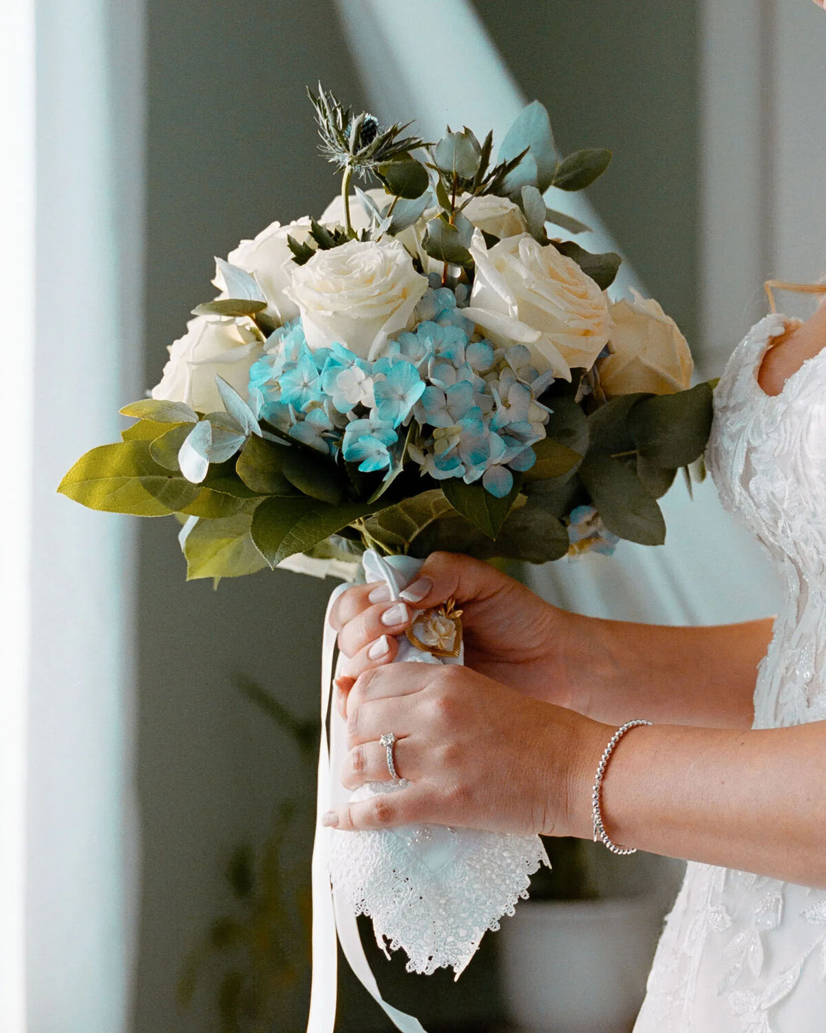 A bride holding a bouquet of white roses, blue hydrangeas, and green leaves. Captured by an NJ wedding photographer, she wears a white dress, a bracelet, and an engagement ring, with a lace handkerchief wrapped around the bouquet stems.