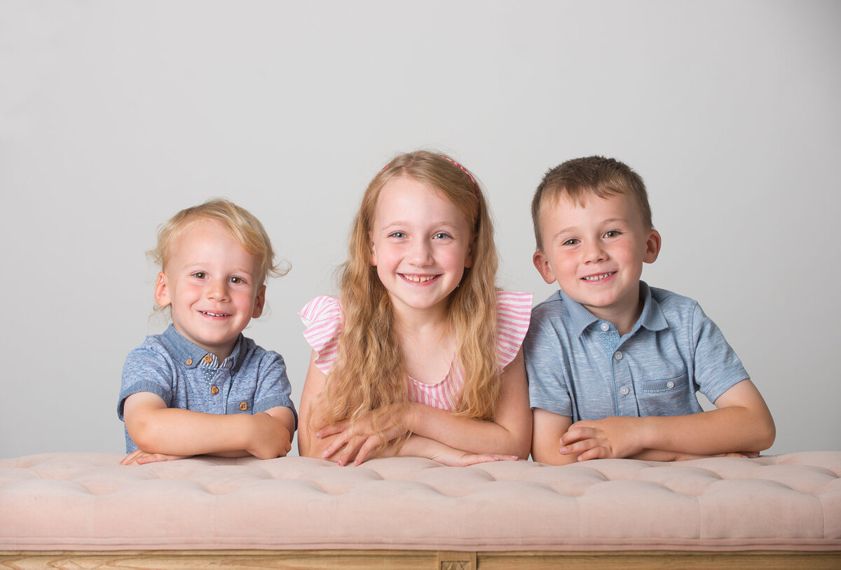 portrait of three children leaning on coral bench smiling wearing blue and coral tops