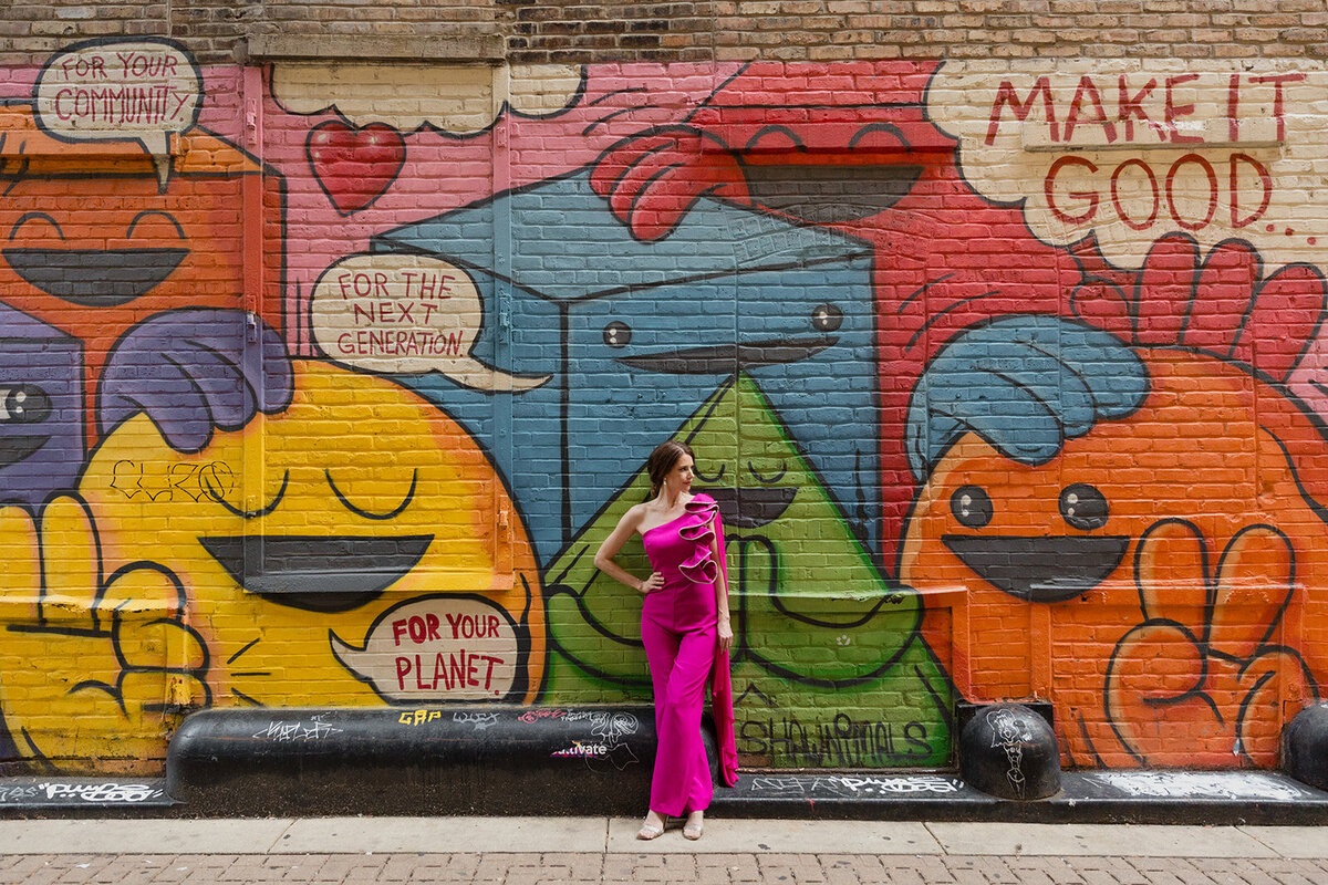 woman is leaning against a colorful mural, wearing a bright pink jumpsuit. She is looking off to the side.