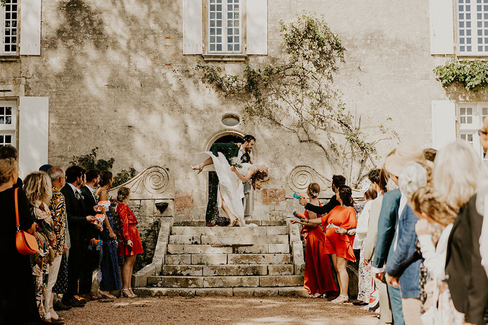 Marié renversant en arrière la mariée sur les marches d'un bâtiment ancien. Haie d'honneur des invités au premier plan. Shooting photo mariage en Vendée.