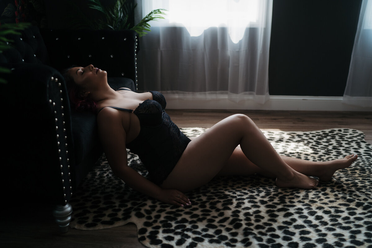 A woman in black lace lingerie sits on a cheetah print rug in front of a window while leaning back onto a black couch