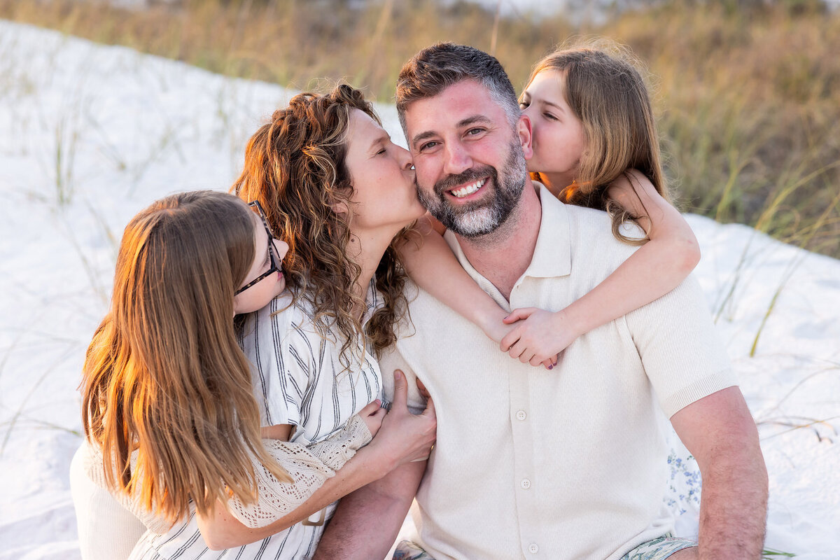 family of 4 sitting on sand in front of Watercolor Inn and the 2 daughters are kissing the dad while mom looks at her husband, Gwyne Gray Photography