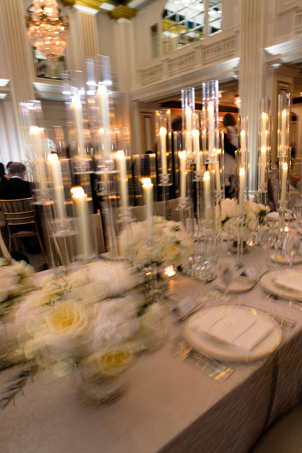 Elegant dining table with tall candle holders and white floral arrangements, set in a luxurious room with chandeliers and high ceilings. The ambiance is warm and inviting, perfect for a sophisticated gathering or event.