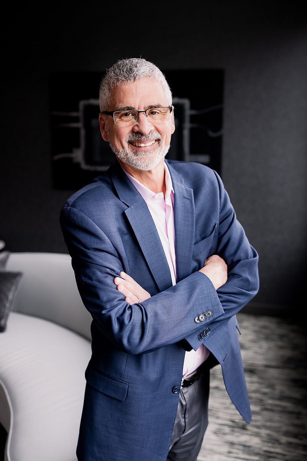 Mark Segal smiling with his arms across his chest in a suit jacket with a dark wall background