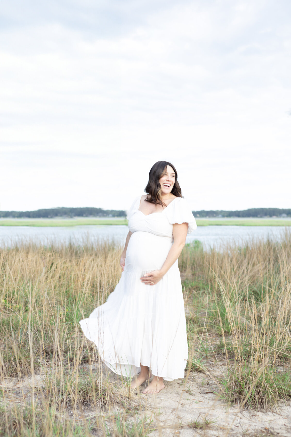 Pregnant woman wearing a white dress standing on the coast, laughing and twirling  by Virginia Beach Maternity Photographer Mary Eleanor Photography