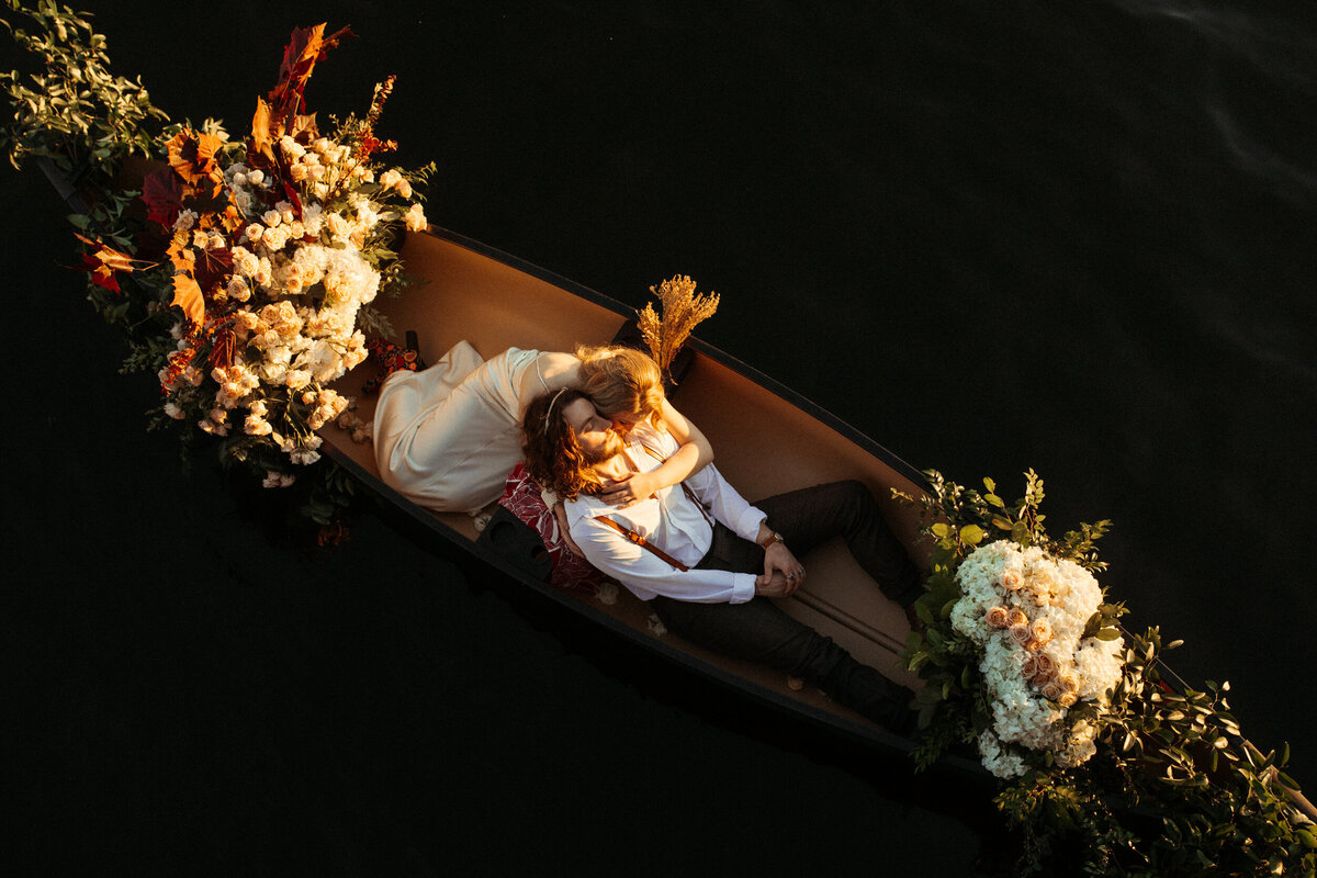 Aerial view of a bride and groom laying in a boat decorated with flowers as they float on a lake at sunset