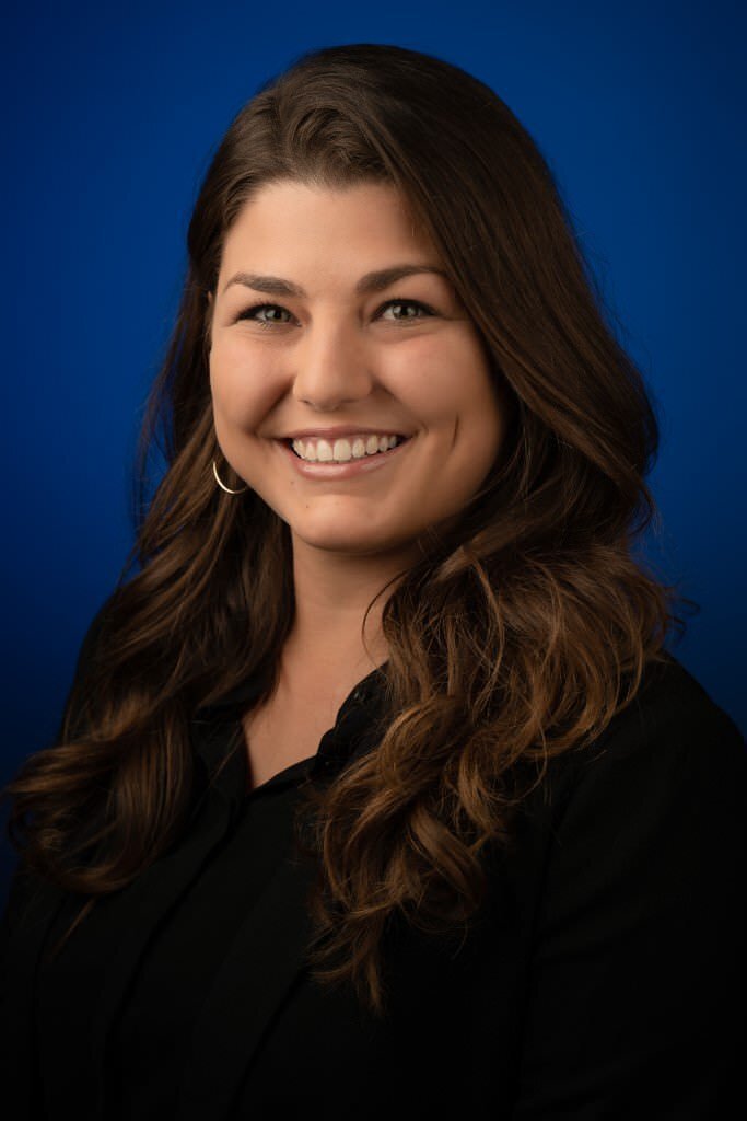A-smiling-woman-with-long-brown-hair-dressed-in-a-black-blouse-against-a-blue-background