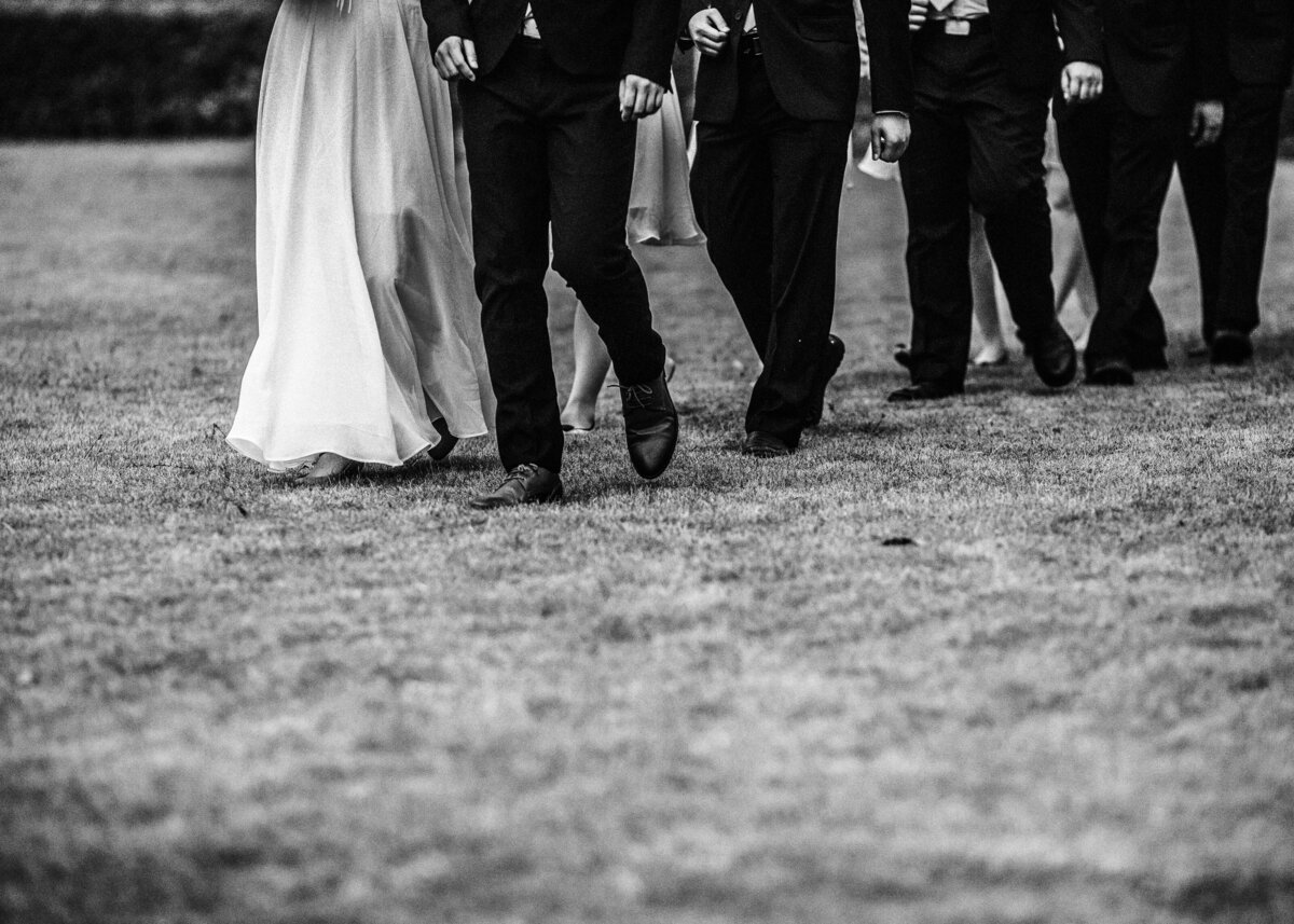 A bride walks with a group of ushers across a grass lawn.