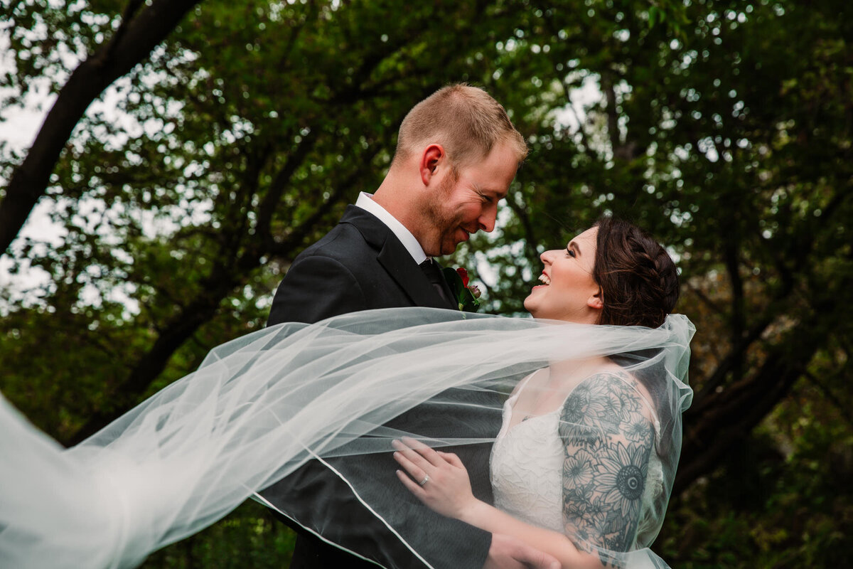 bride and groom smile at each other while veil blows foward towards groom