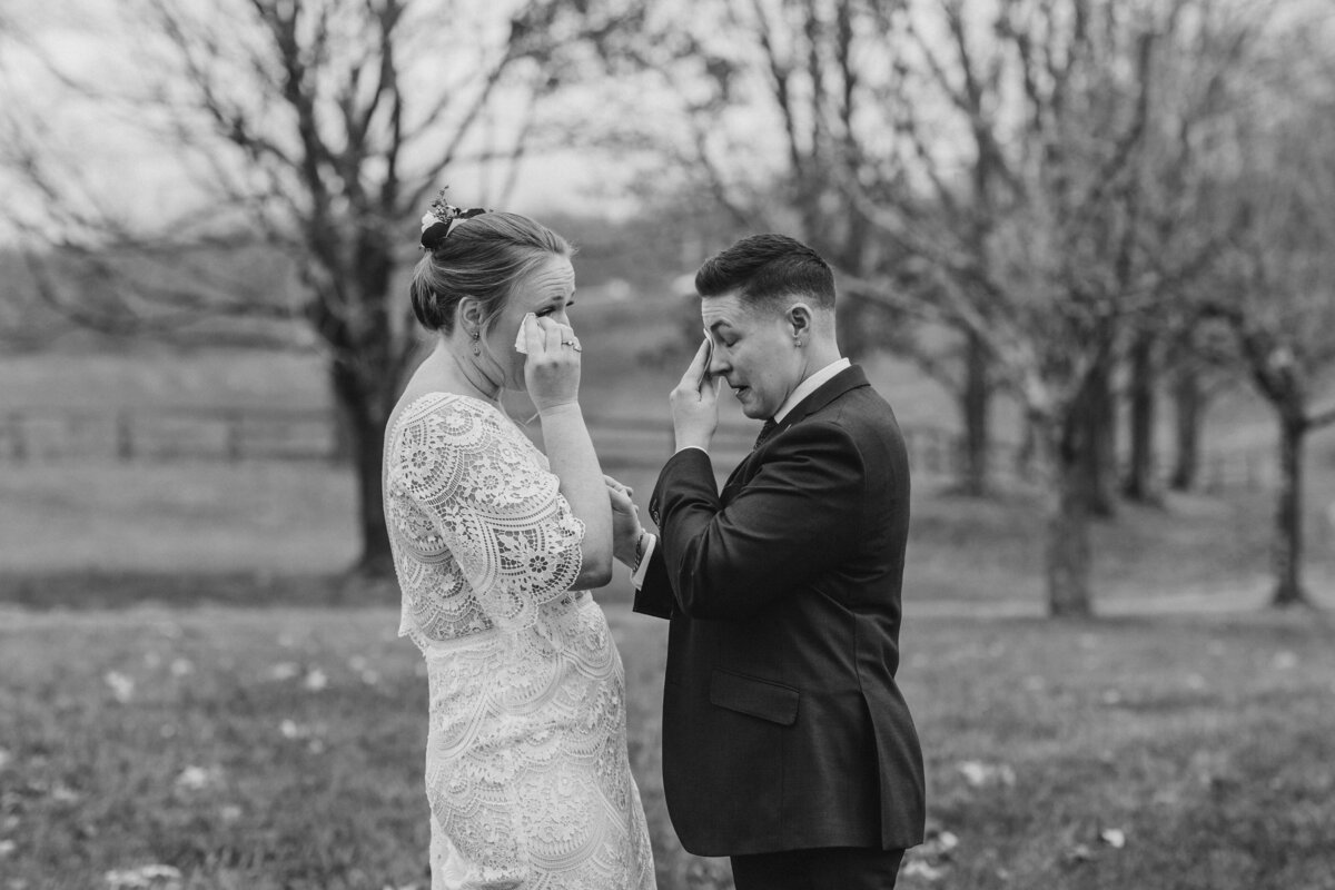 A newlywed couple standing close as they wipe their own tears 