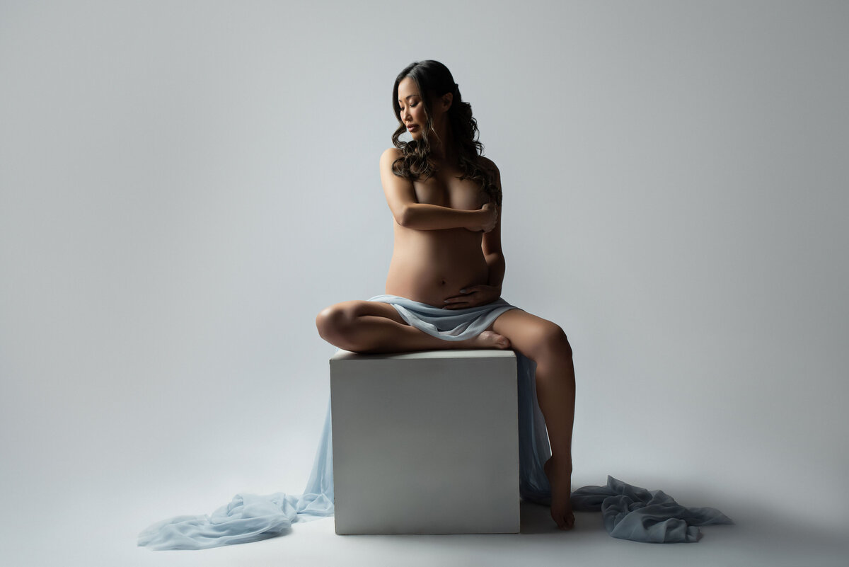 32 weeks pregnant asian women sitting on studio posing cube with draped fabric across her legs with hands covering her chest and looking into the distance  gorgeous studio maternity
