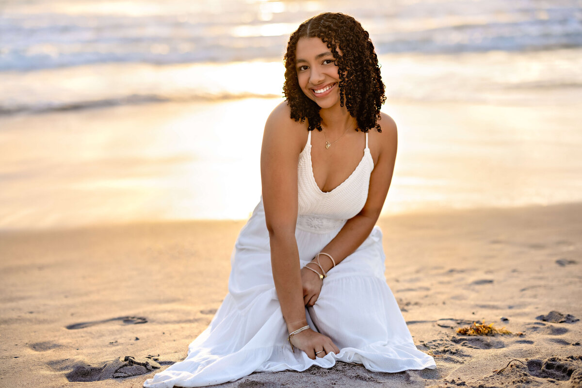 Winter Garden senior portraits with a girl sitting in the sand at sunrise, leaning forward with a natural smile and glowing backlight.