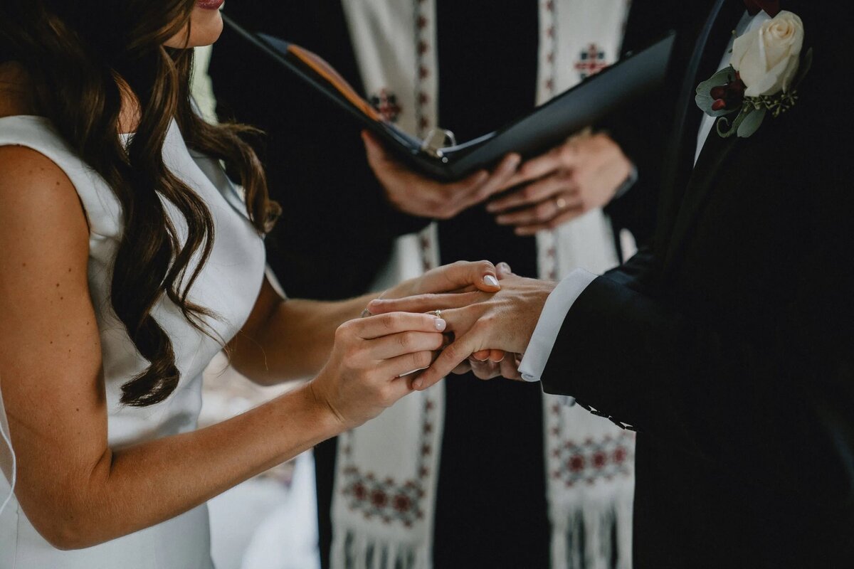 A close-up of a bride and groom's hands during their wedding ceremony, with the groom placing a ring on the bride's finger.