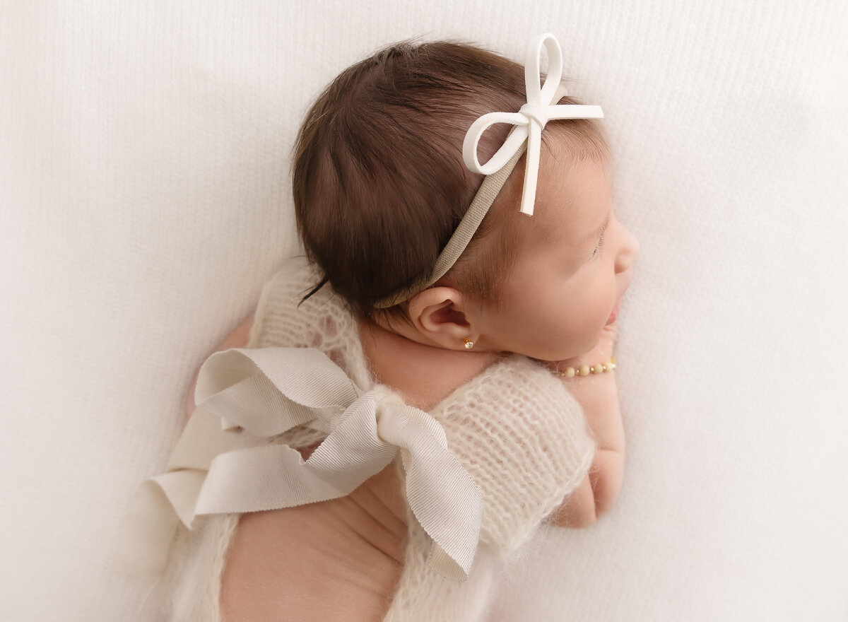 Baby girl is laying on her belly - aerial image features baby's back and face side profile. Baby is wearing a cream knit romper with large bow at the back. Captured by top Brooklyn newborn photographer Chaya Bornstein