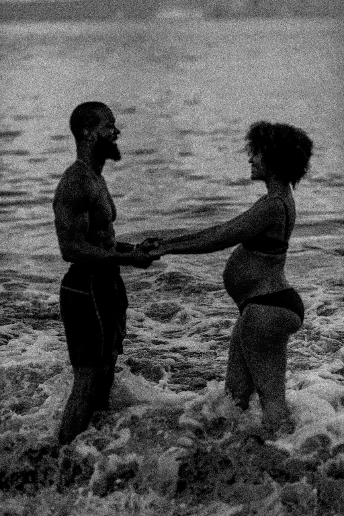 Black and white image of a couple holding hands in the ocean during babymoon photography session in Hawaii with Love + Water