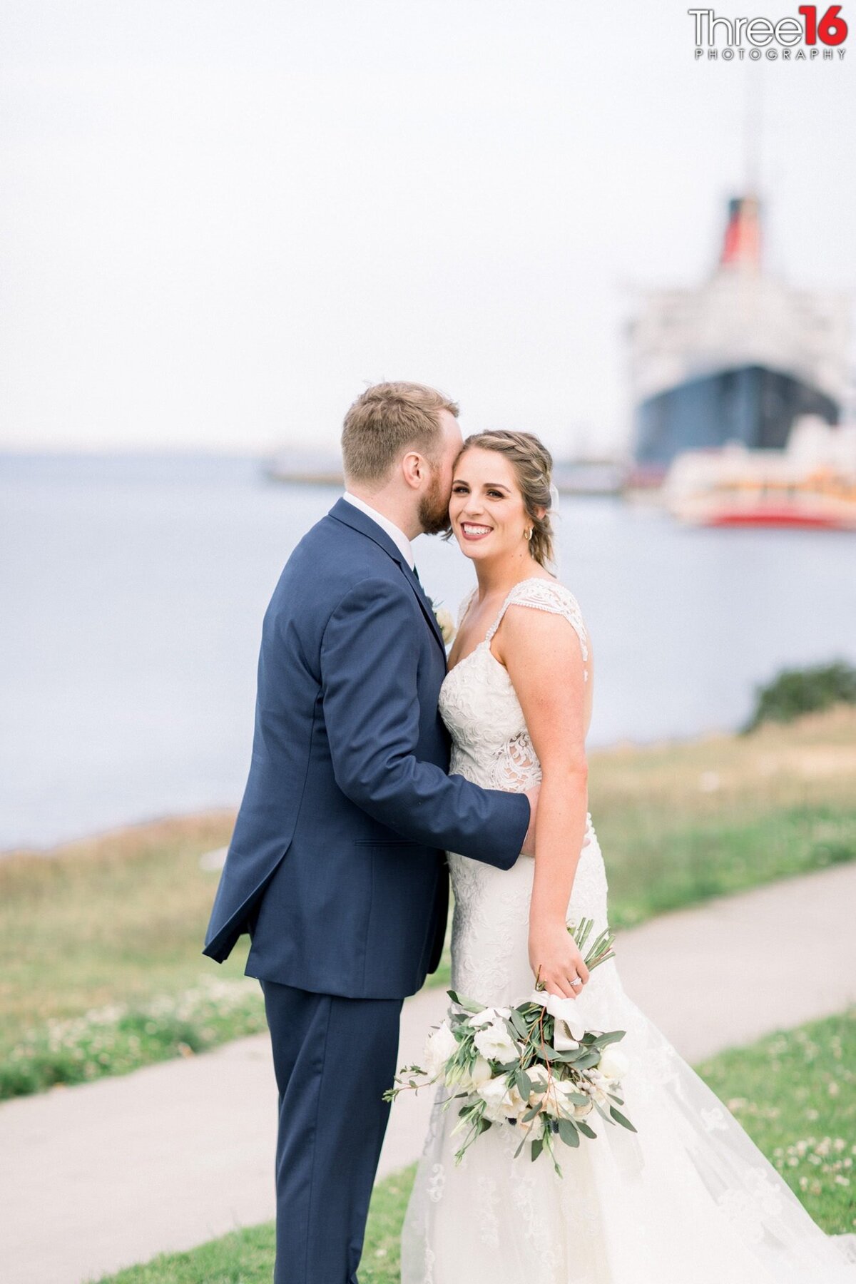 Groom embraces his Bride as she smiles at the wedding photographer with the Queen Mary in the background