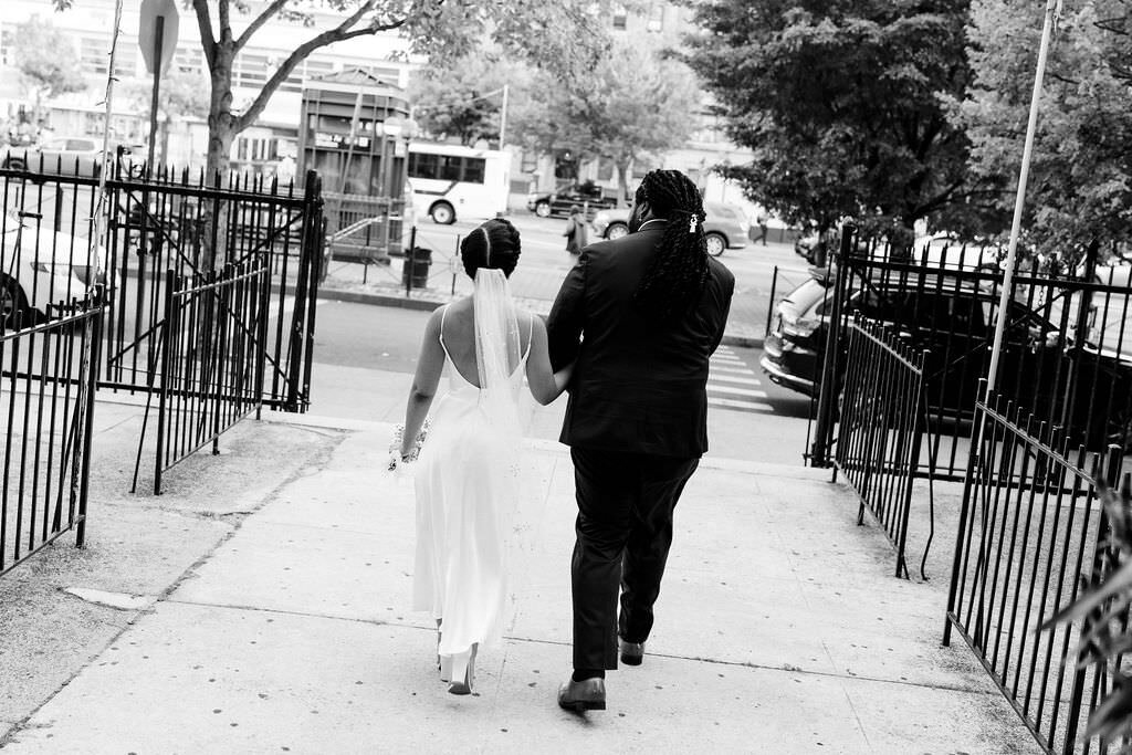 black and white photo of a bride and groom walking along a sidewalk through a gate