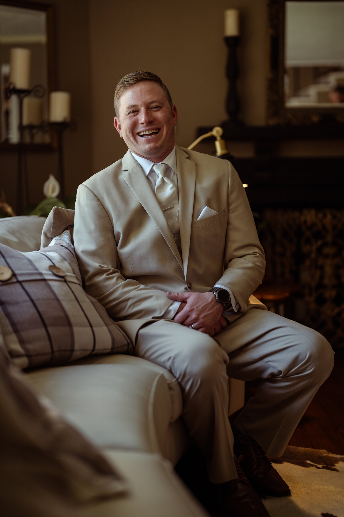 Candid portrait of a groom smiling warmly while sitting on a couch on his wedding day. Dressed in a beige suit with a matching tie and pocket square, he exudes a relaxed and joyful vibe, captured in an elegantly decorated indoor setting.