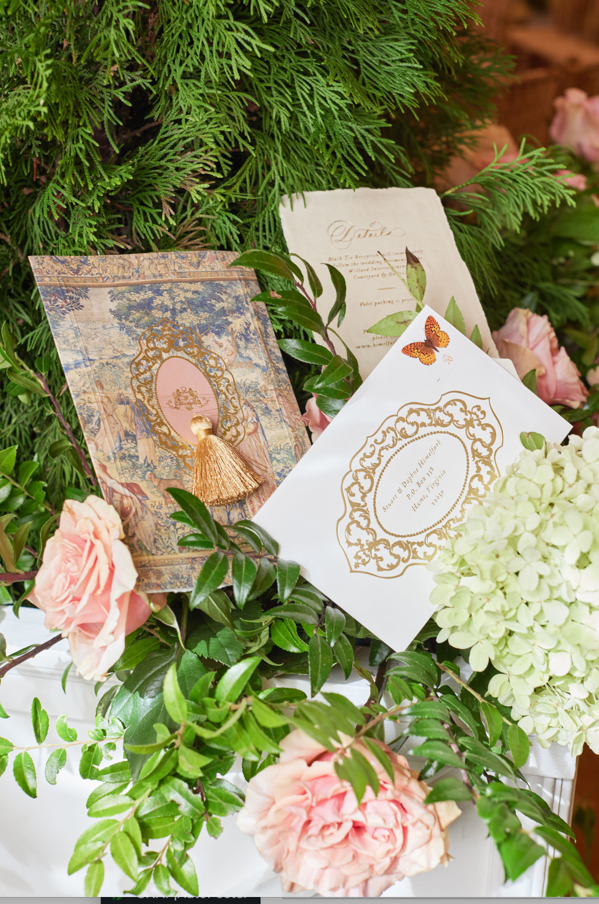 The photo was taken at Washington D.C., The Willard Intercontinental, one of the top wedding venues and hotels in DC by photographer Shawn Connell. The scene captures a set of intricately designed greeting cards displayed amid lush greenery and pink roses. The cards feature ornate patterns, tassels, and one with a butterfly, adding a touch of whimsy to the elegant display. A hydrangea bloom and leaves are also incorporated, seamlessly blending nature with the ornamental arrangement.
