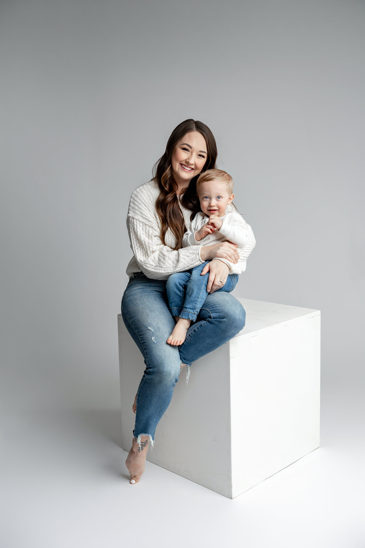 Mother sitting on white cube holding her young son in denim and neutral outfits. Photographed by Yucaipa family photographer Kaitlyn Dawn Photography.