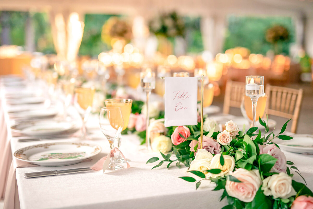 A beautifully set wedding reception table with elegant floral arrangements of pink and white roses. Crystal glassware and plates are neatly arranged. A sign reading Table One is visible. Warm lighting creates a soft, inviting atmosphere.