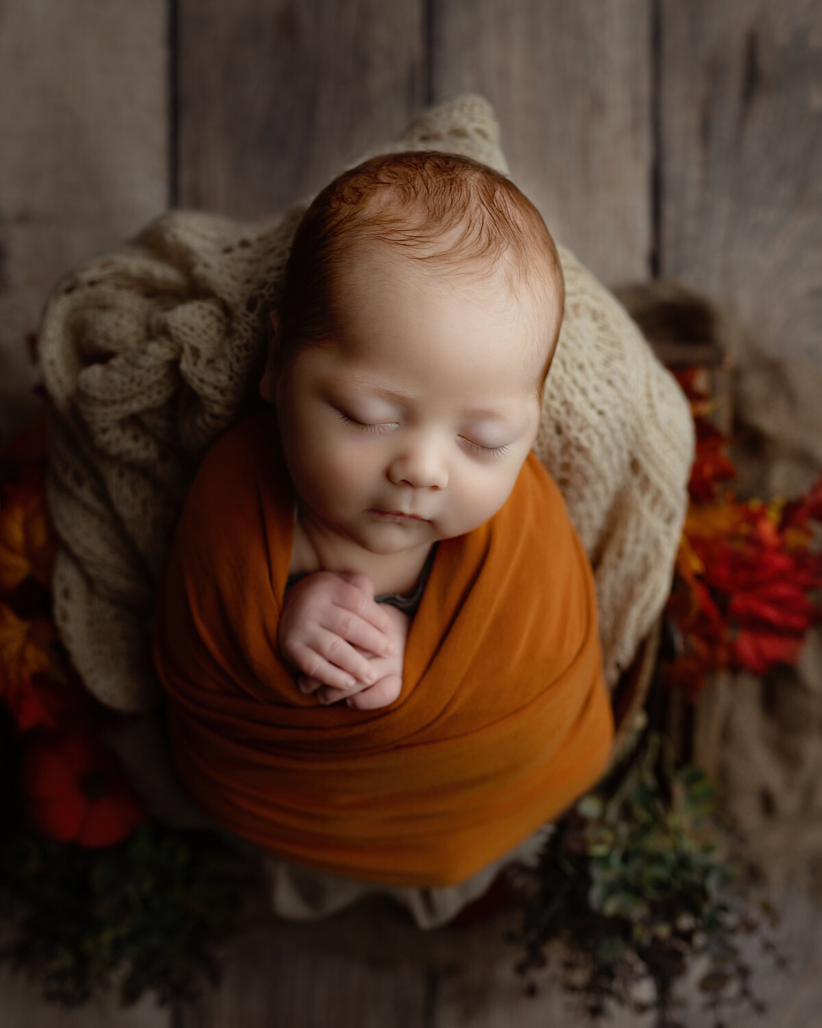 A serene moment captured close-up—baby wrapped in burnt orange with delicate hands and surrounded by cozy fall tones.