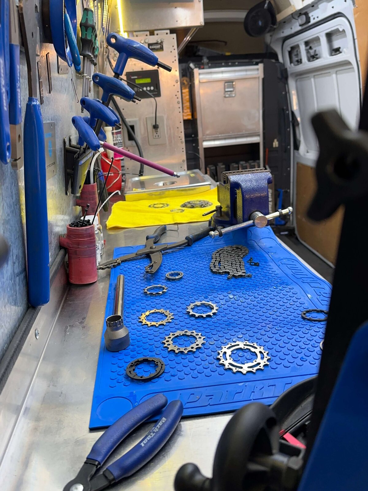 A bicycle repair workspace with tools and bike parts. Blue mats, chainrings, and chains are organized on a metal bench. Pliers and various tools are visible, along with storage drawers in the background