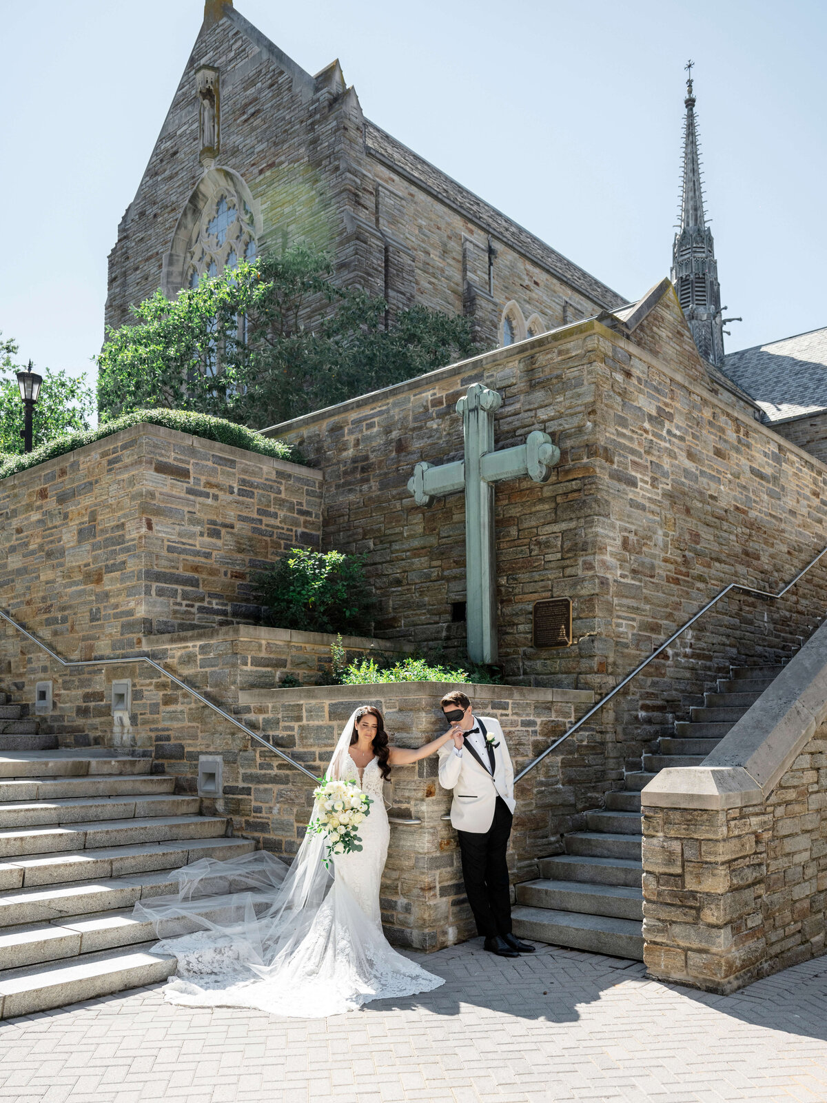 A bride in a white wedding dress and a groom in a white tuxedo stand on stone steps outside a large stone church with a cross. The bride holds a bouquet, and both look relaxed in the sunny setting.