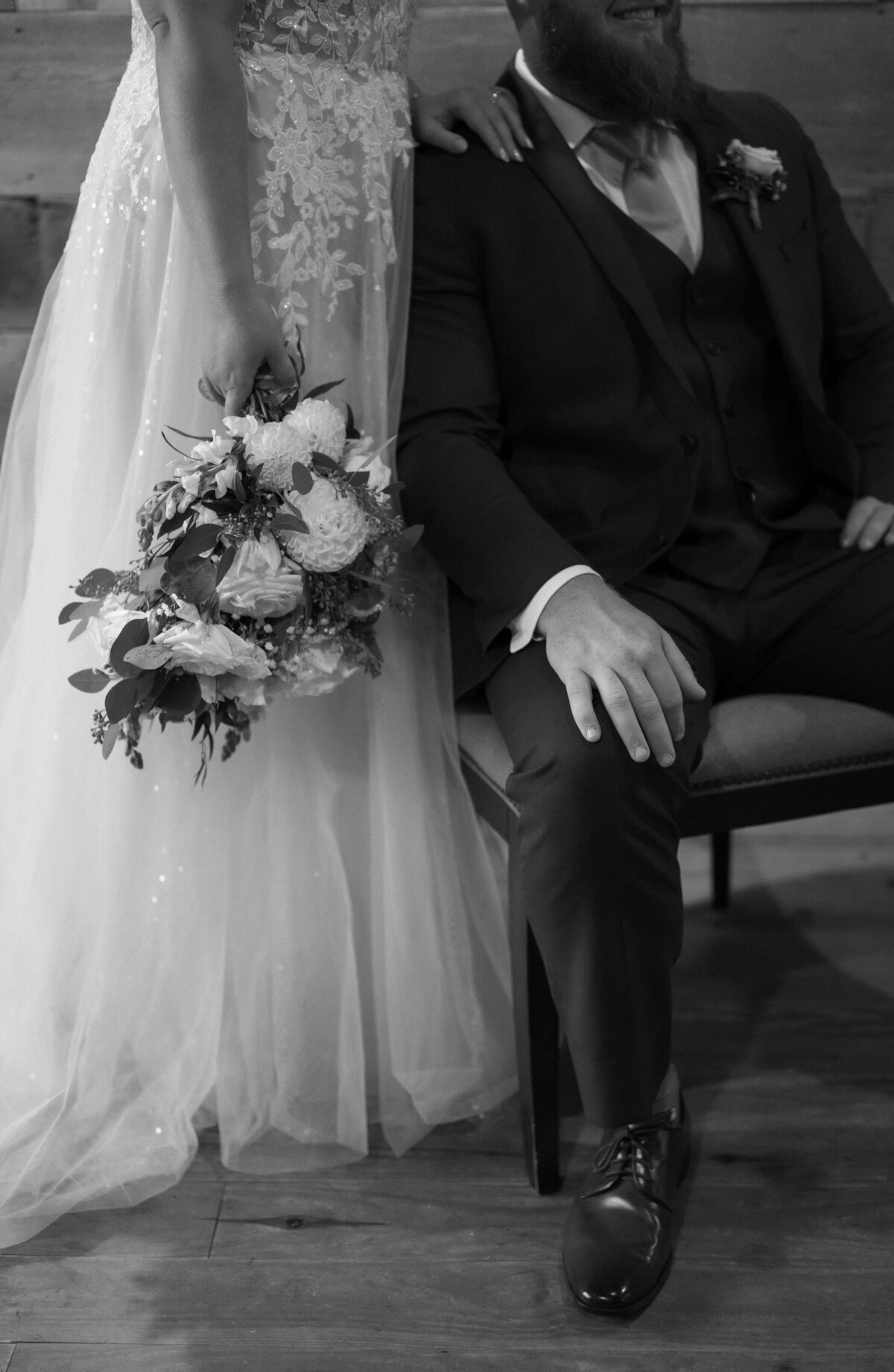 black and white wedding photo for a Shenandoah national park wedding with groom sitting on a chair in a living room with his bride standing to his side and holds her bouquet to her side